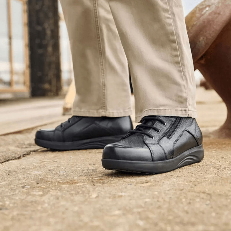 Ein Mann in beiger Hose und schwarzen New Feet Herrenstiefeln steht auf grauem Untergrund